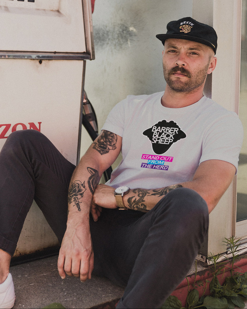 Man sitting on a step wearing a t-shirt with a graphic design, in front of a gas pump.