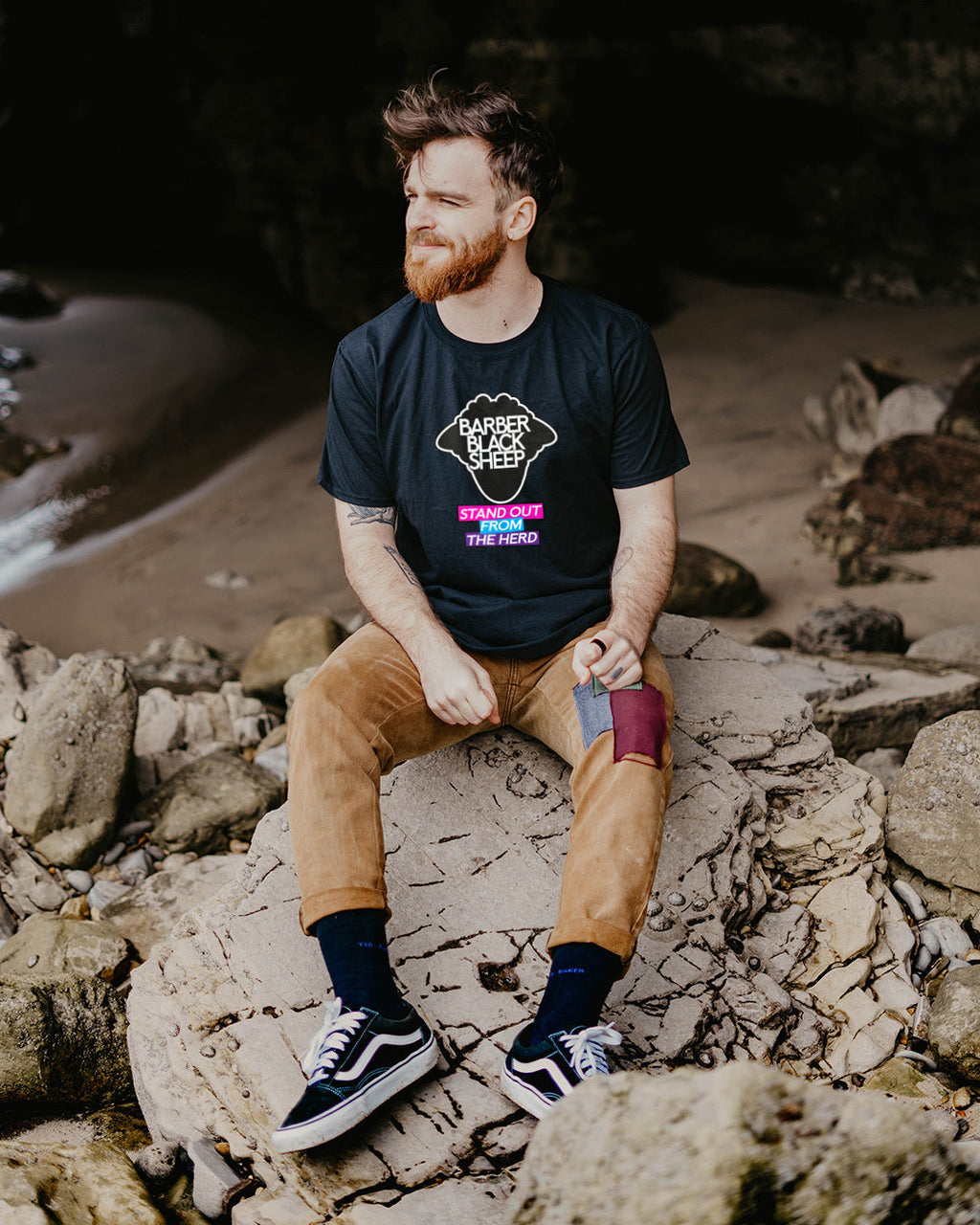 Man sitting on rocks by a cave entrance wearing a black t-shirt with colorful graphics.
