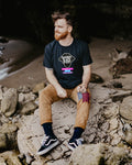 Man sitting on rocks by a cave entrance wearing a black t-shirt with colorful graphics.