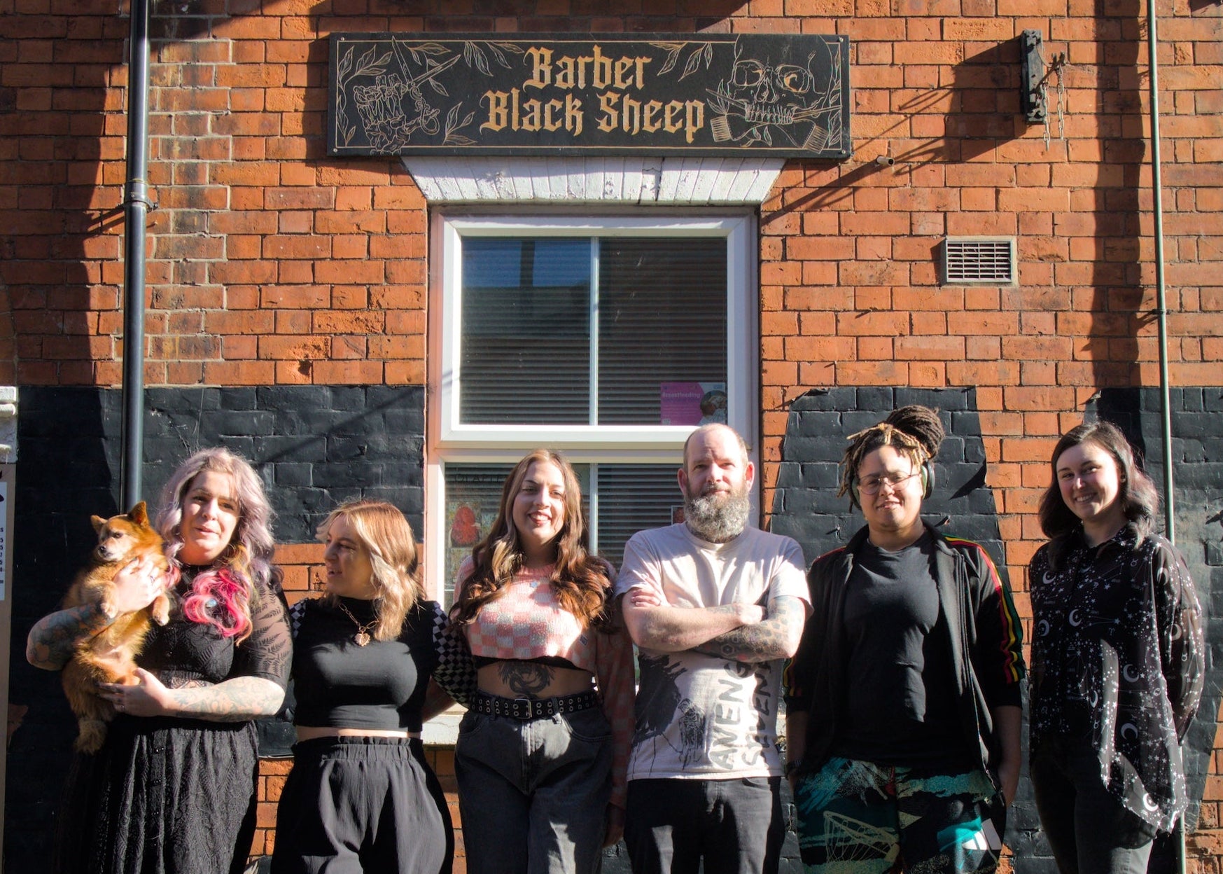 Group of people standing in front of a brick building with a sign that reads 'Barber Black Sheep'.